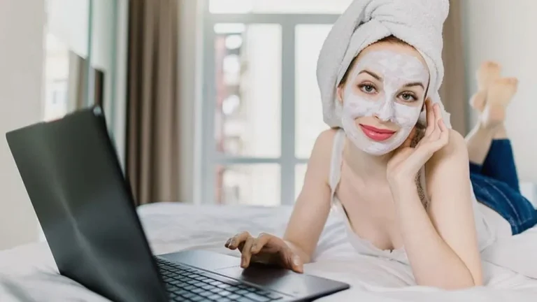 A woman with a face mask and hair towel using a laptop on a bed, illustrating the relaxed lifestyle of how to remove gel nails at home.
