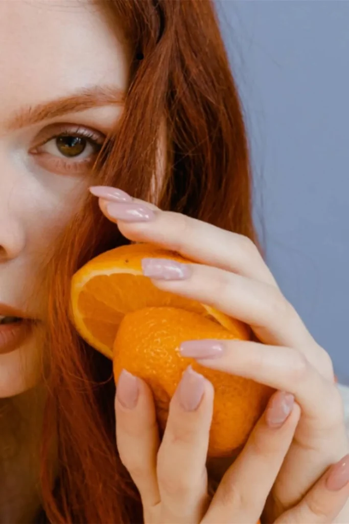 Close-up of a redhead woman holding an orange to show the importance of Vitamin C in learning how to grow nails fast.
