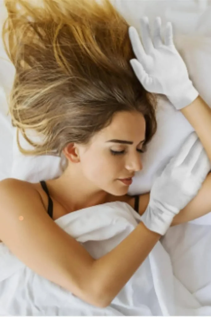 A woman sleeping in bed wearing white cotton gloves to demonstrate an overnight hydration routine for how to grow nails fast.